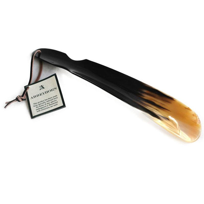 A flat-shaped shoe horn with a cut-out handle and a leather thong strap, made of natural horn, with a product label attached.
