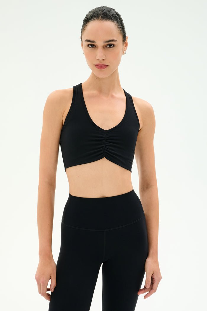 A woman models the SPLITS59 Airweight Bra with Ruching in black, ideal for hot yoga, paired with high-waisted leggings against a plain white background.