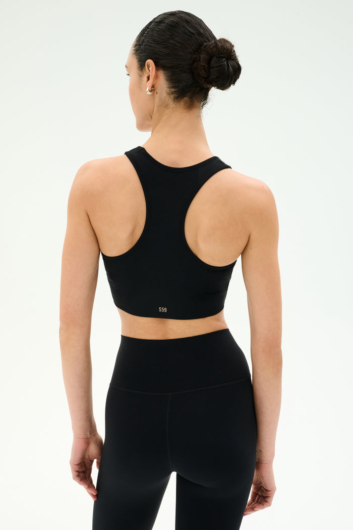 A woman faces away in the SPLITS59 Airweight Bra with Ruching - Black, paired with black high-waisted leggings against a plain light background—ideal for hot yoga sessions.