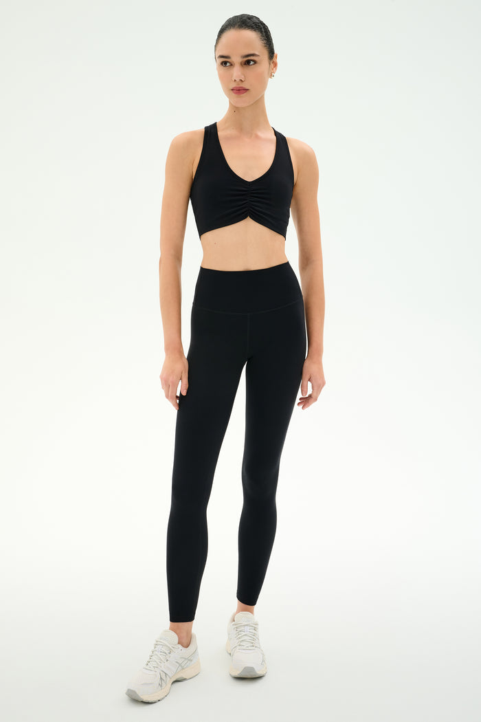 Model wearing SPLITS59 Airweight Bra with Ruching in black, paired with high-waisted black leggings and white athletic shoes, poses against a white background—ideal activewear for hot yoga sessions.