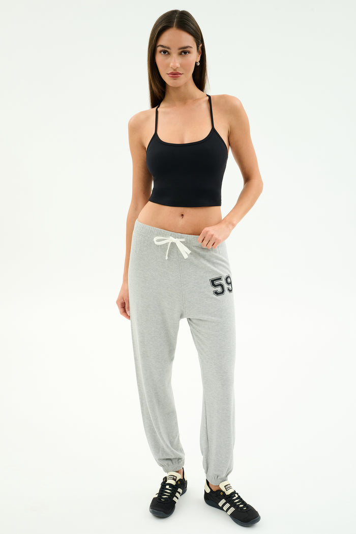 A woman stands against a white background wearing a black SPLITS59 Airweight Crop, light gray sweatpants with "59" on the left side, and black sneakers—ideal for hot yoga or relaxed casual days.