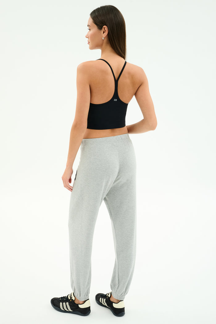 A woman faces away, wearing the SPLITS59 Airweight Crop in black, paired with grey sweatpants and black sneakers with white stripes, posed against a plain white background.