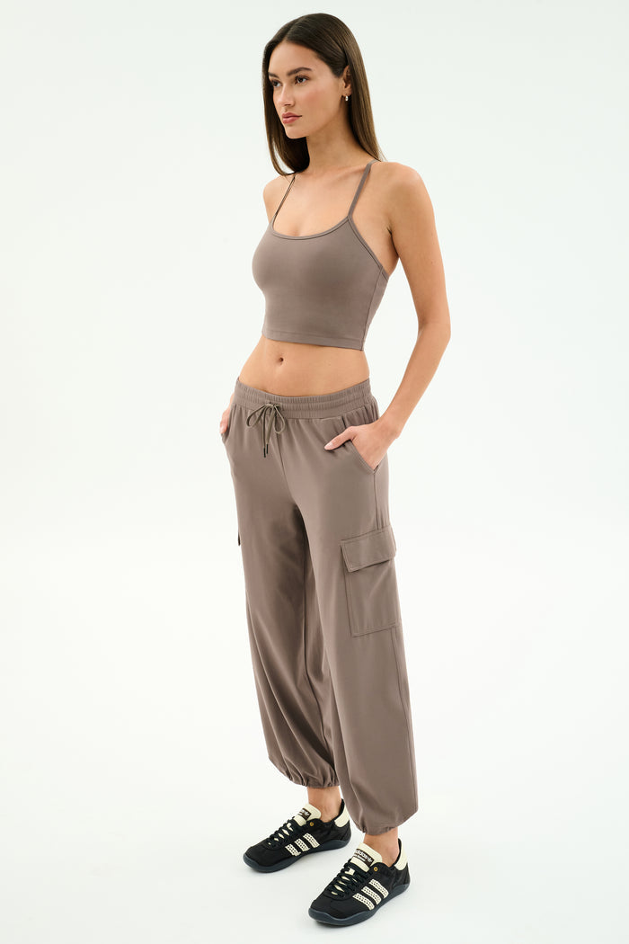 A woman poses against a white background in the SPLITS59 Airweight Crop - Lentil and matching cargo pants, paired with black sneakers with white stripes. Her hands in her pockets showcase an effortlessly cool look for hot yoga or casual outings.