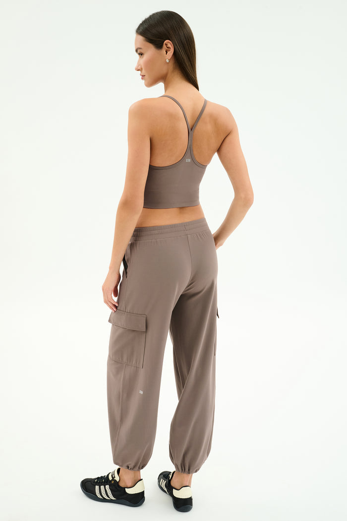 A woman faces away, wearing the SPLITS59 Airweight Crop in Lentil and matching joggers with black Adidas sneakers against a plain light background.