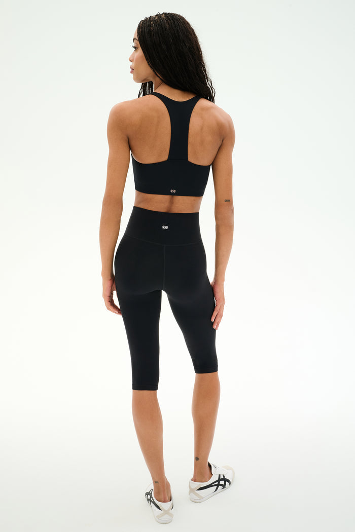 A woman stands on a white background, viewed from the back, wearing the SPLITS59 Airweight High Waist 16" Capri Legging in black, a black sports bra, and white sneakers.