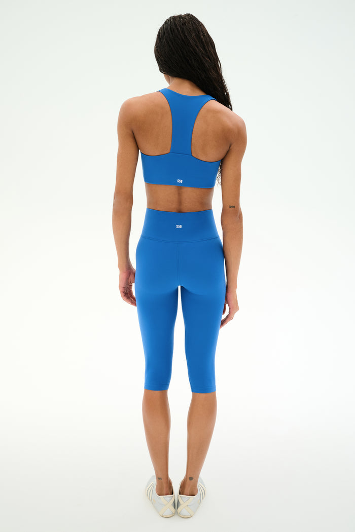 A person stands with their back to the camera, wearing the SPLITS59 Airweight High Waist 16" Capri Legging in Classic Blue, paired with a matching blue sports bra, against a plain white background.