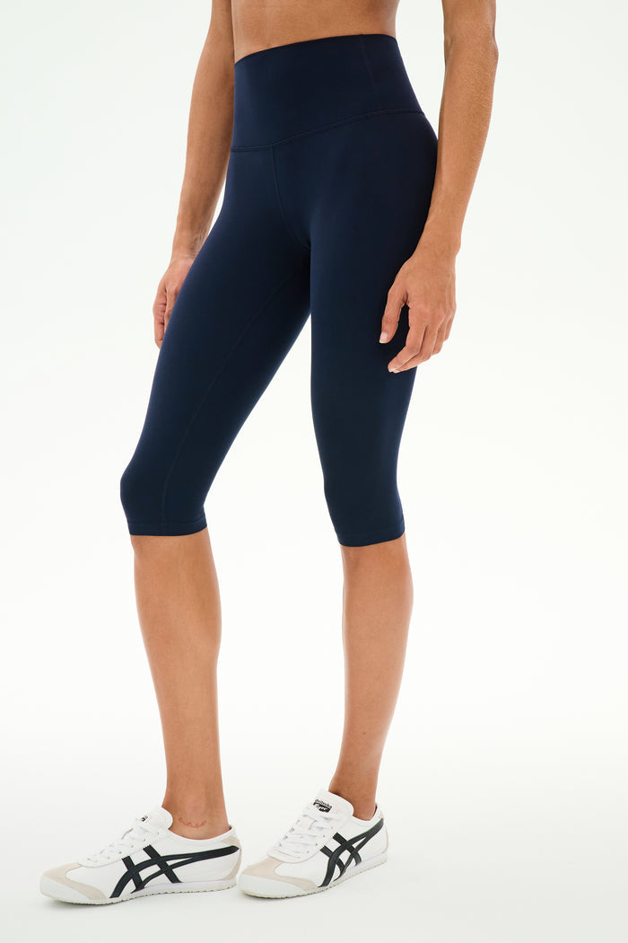 A person wearing the SPLITS59 Airweight High Waist 16" Capri Legging in Indigo and white sneakers, standing against a plain white background.