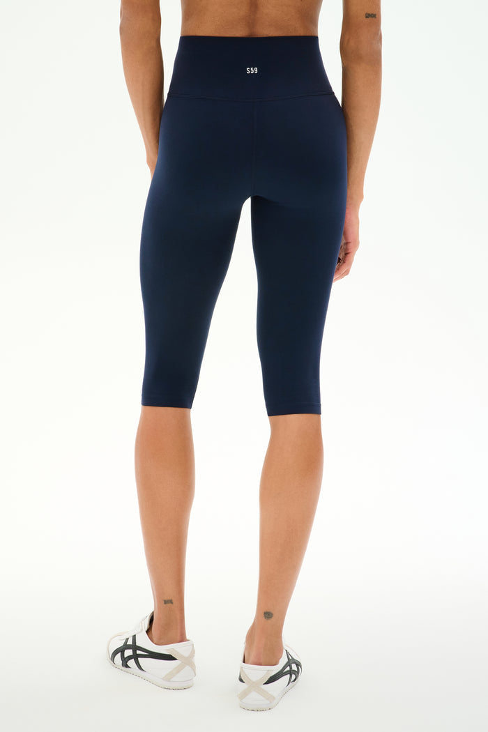 A person is wearing SPLITS59 Airweight High Waist 16" Capri Legging in Indigo and white sneakers, standing on a white background, viewed from the back.