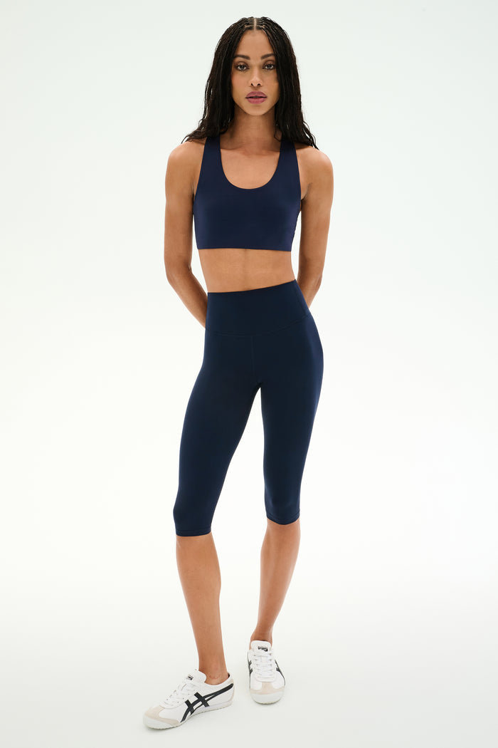 A woman stands against a white background, wearing the SPLITS59 Airweight High Waist 16" Capri Legging in Indigo, a navy blue sports bra, and white sneakers.