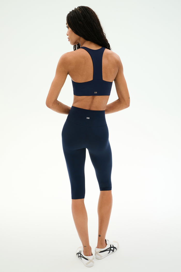 A person is shown from the back, standing on a white background, wearing a navy blue high-waisted sports bra and matching SPLITS59 Airweight High Waist 16" Capri Legging in Indigo with white sneakers.