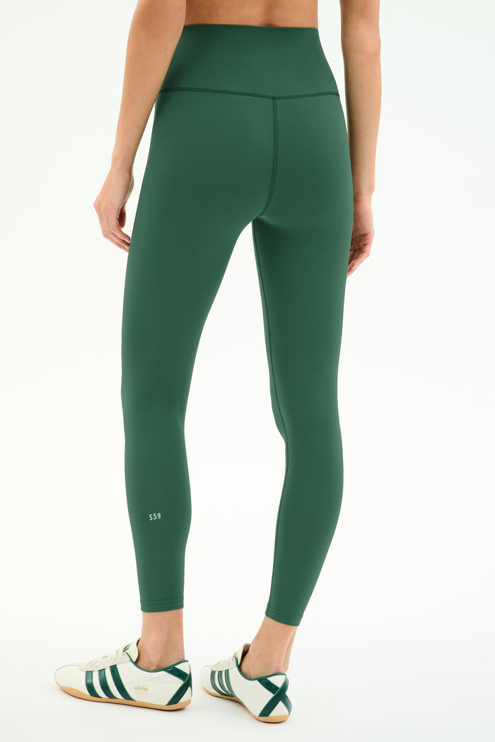 A person is seen from the back wearing SPLITS59 Airweight High Waist Legging in Arugula and white sneakers with green stripes, standing against a plain white background—ideal for hot yoga sessions.