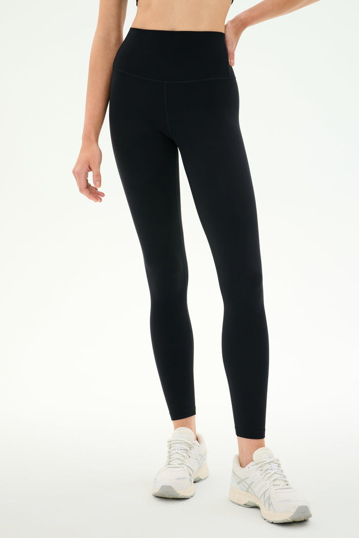 A person wearing SPLITS59 Airweight High Waist Legging in black and white athletic shoes stands against a plain light background, ready for hot yoga.