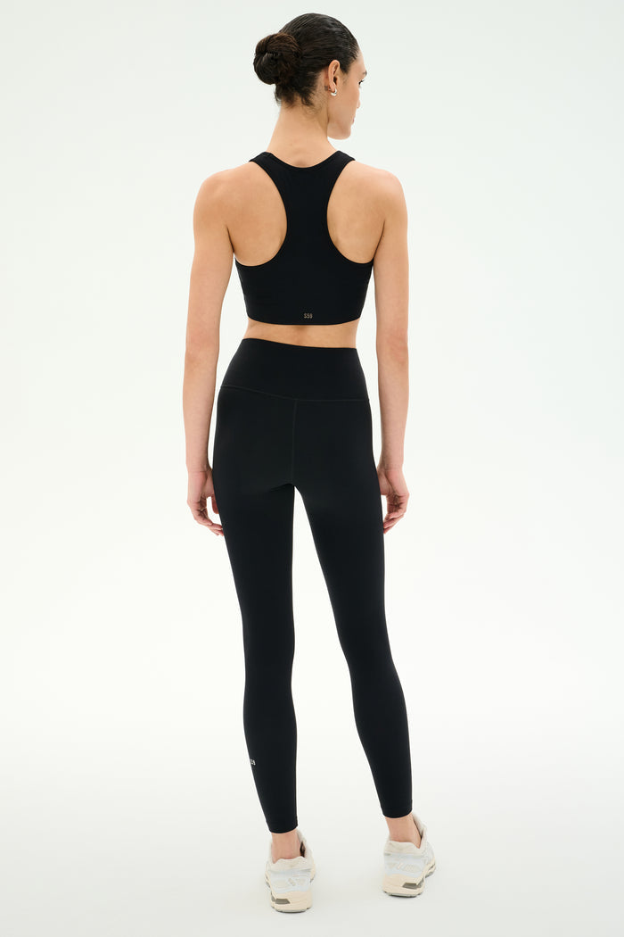 A woman faces away wearing SPLITS59's Airweight High Waist Legging in black with a matching sports bra and white sneakers against a plain, light background—ideal for hot yoga sessions.