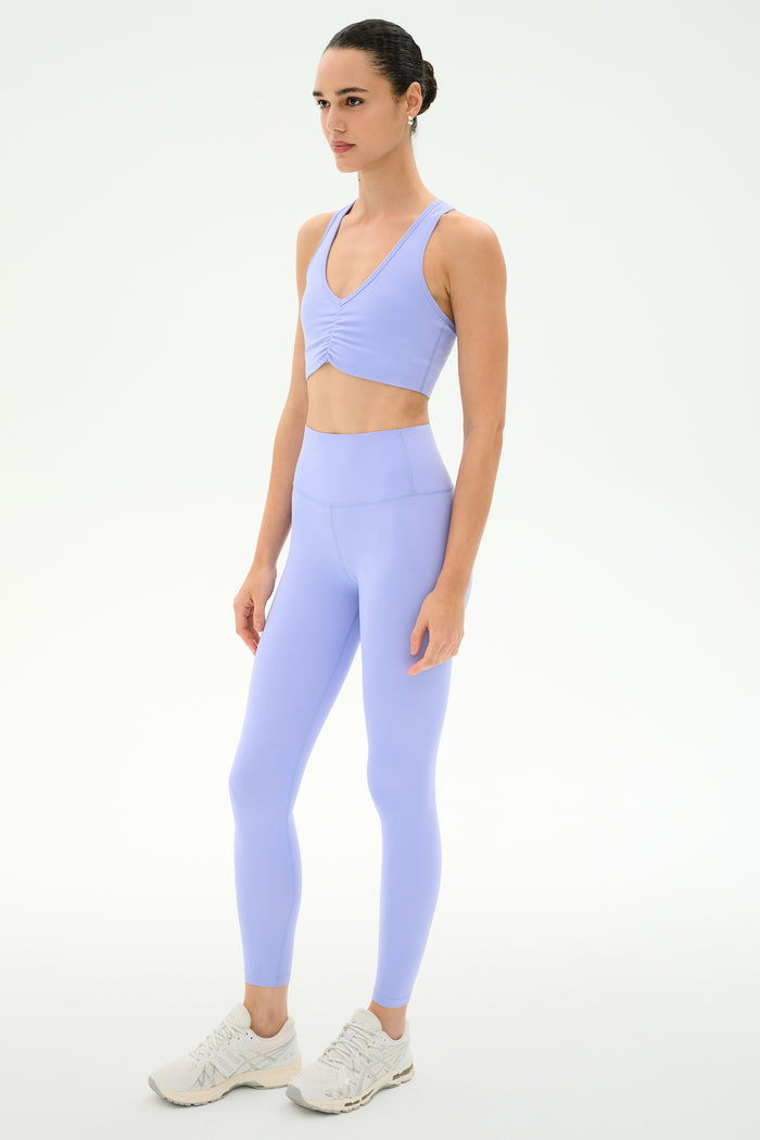 A woman stands against a plain white background, wearing the SPLITS59 Airweight High Waist Legging in Purple Haze with a matching light purple sports bra and white athletic shoes—ultra-soft for hot yoga or your next studio session.