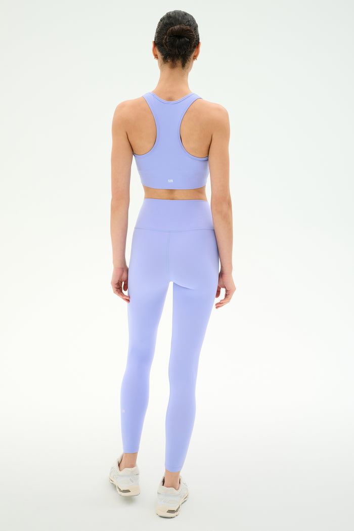 A person stands facing away on a plain white background, wearing the SPLITS59 Airweight High Waist Legging in Purple Haze with a matching light purple sports bra and white sneakers.