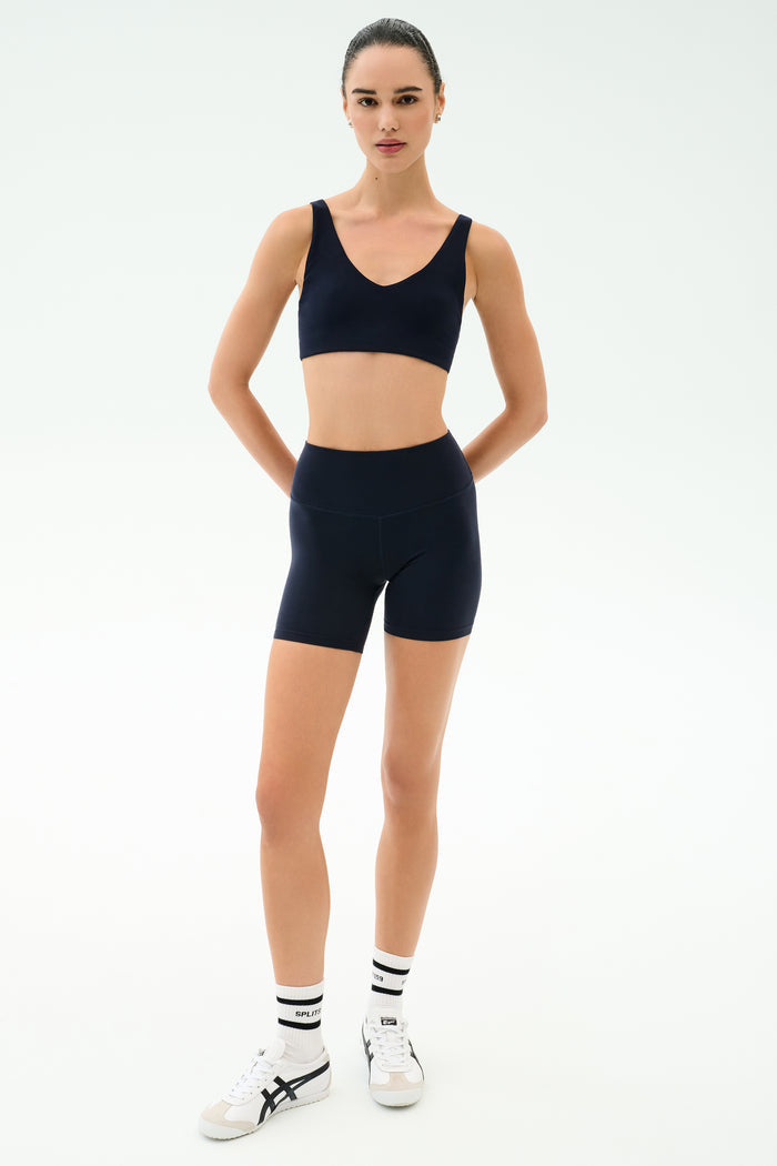 A woman faces forward wearing the SPLITS59 Airweight High Waist Short in Indigo, a matching navy seamless tank with a shelf bra, white socks with black stripes, and white athletic shoes against a plain light background.
