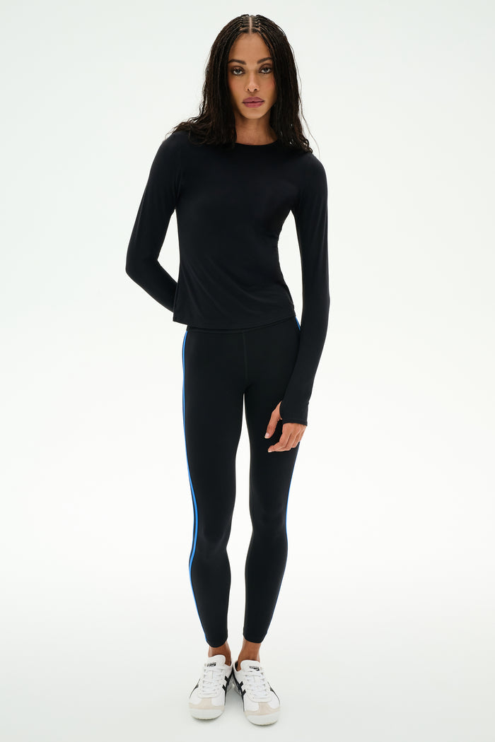 A woman stands against a plain background wearing the SPLITS59 Airweight Lite Long Sleeve Waist Length top in black—perfect attire for quick dry comfort during gym workouts.