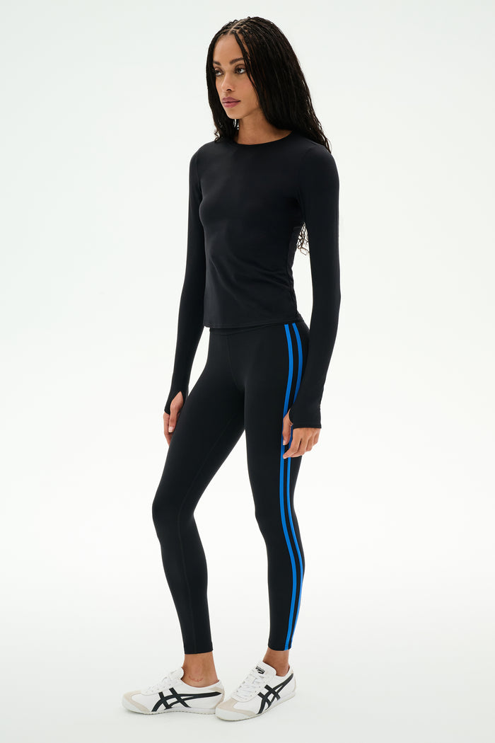 A woman is standing against a plain white background, dressed in the SPLITS59 Airweight Lite Long Sleeve Waist Length top in black, paired with black leggings featuring blue side stripes and white sneakers—an ideal outfit for gym workouts.