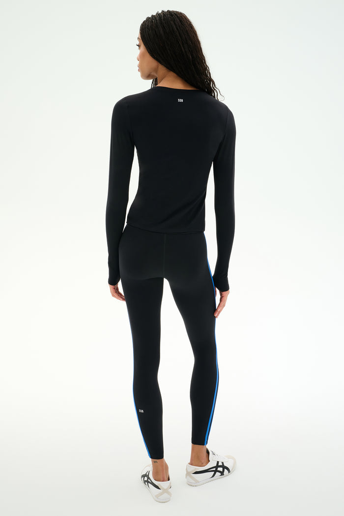 A person stands facing away, wearing the SPLITS59 Airweight Lite Long Sleeve Waist Length top in black and matching leggings with blue side stripes, paired with white sneakers against a plain light background—an ideal outfit for gym workouts.