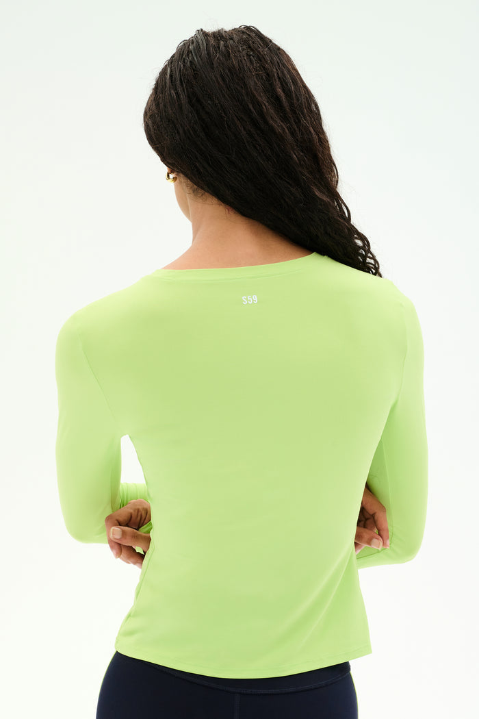 A person with long dark hair, seen from behind, wears the SPLITS59 Airweight Lite Long Sleeve Waist Length top in Limeade paired with dark pants—an ideal gym workout look—set against a plain white background.