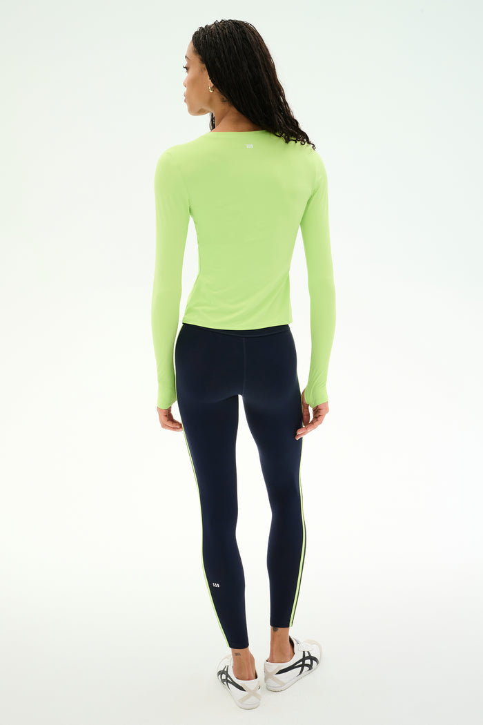 A woman stands with her back to the camera, modeling the SPLITS59 Airweight Lite Long Sleeve Waist Length tee in Limeade, a lightweight, waist-length top with long sleeves. She pairs it with dark leggings featuring side stripes and white sneakers, creating a comfortable gym-ready outfit against a plain white backdrop.