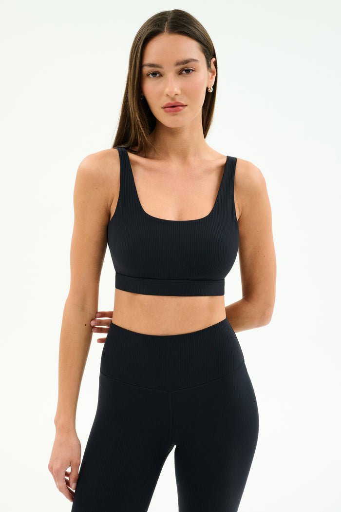 A woman models the SPLITS59 Airweight Rib Bra in black with high-waisted leggings against a plain white background.