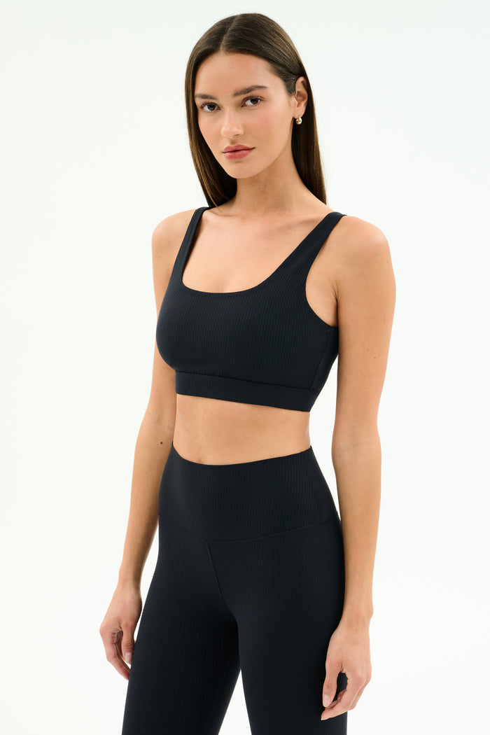 A woman with straight brown hair wears the SPLITS59 Airweight Rib Bra in black, paired with high-waisted leggings, both made from moisture-wicking fabric, standing against a plain white background.
