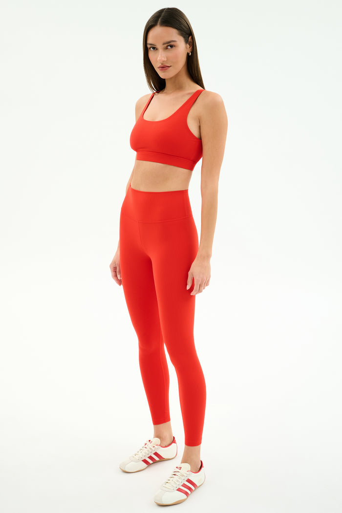A woman poses against a plain background in the SPLITS59 Airweight Rib Bra - Lotus Red, paired with matching leggings and white sneakers with red stripes—an ideal set for hot yoga.