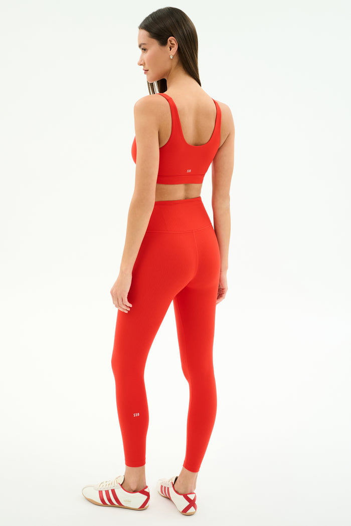 A woman faces away, wearing the SPLITS59 Airweight Rib Bra in Lotus Red with matching leggings and white Adidas sneakers with red stripes, set against a plain white background.