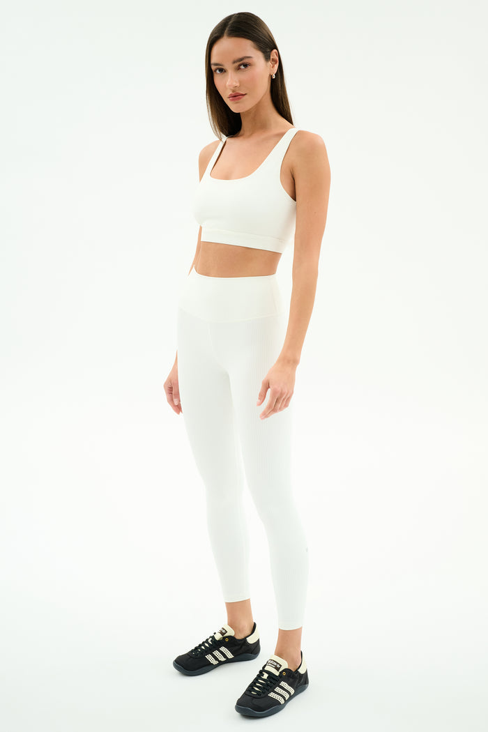 A woman wears the SPLITS59 Airweight Rib High Waist 7/8 leggings in white with a matching sports bra and black sneakers, standing against a plain white background.