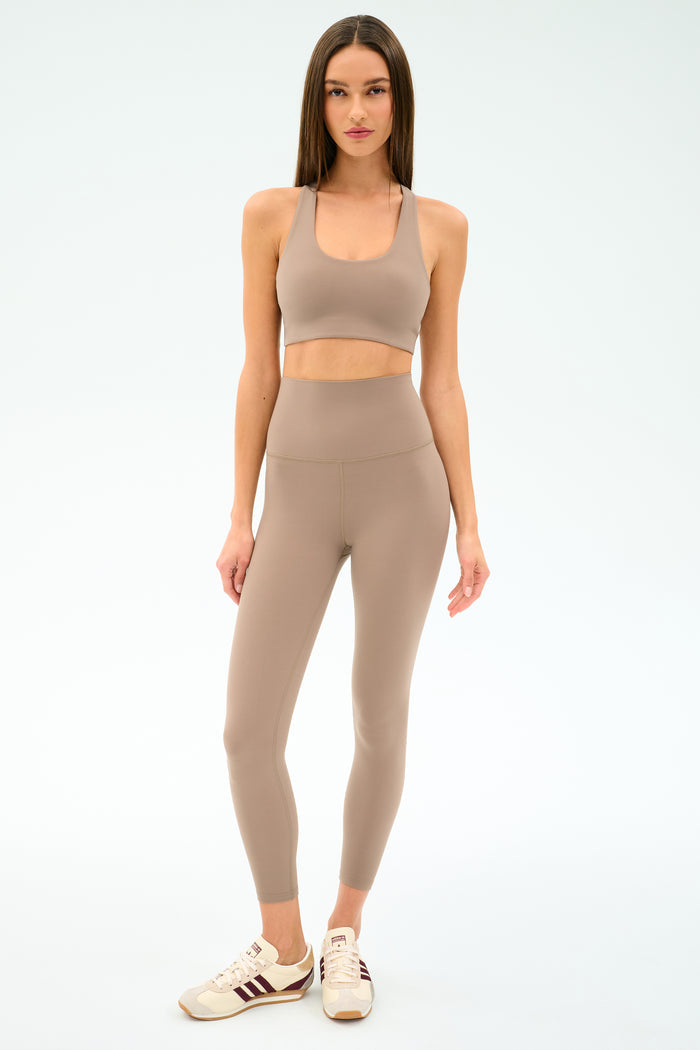 A woman faces forward wearing SPLITS59's Airweight Super High Waist 7/8 leggings in Latte with a matching sports bra—crafted from soft, breathable Airweight fabric—paired with white sneakers against a plain light background.