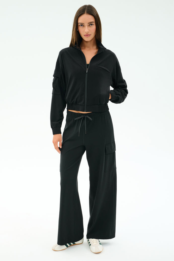 A woman faces forward in a black zip-up jacket and SPLITS59 Alex Airweight Cargo Pant - Black, featuring pockets, paired with white sneakers against a plain white background.