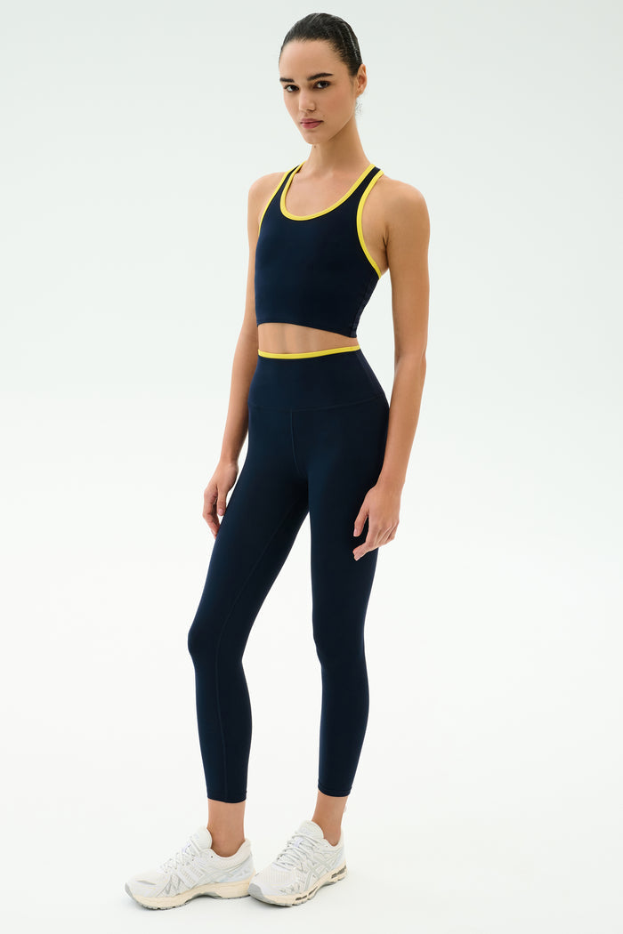 A woman wears the SPLITS59 Amber Airweight Bralette in Indigo/Ray with matching leggings and white sneakers, all crafted from Airweight performance fabric—ideal for hot yoga—posed against a plain white background.