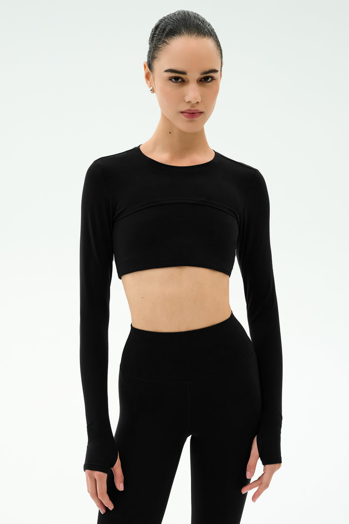 A woman stands against a plain white background wearing the SPLITS59 Amelia Airweight Lite Shrug in black with matching high-waisted leggings.