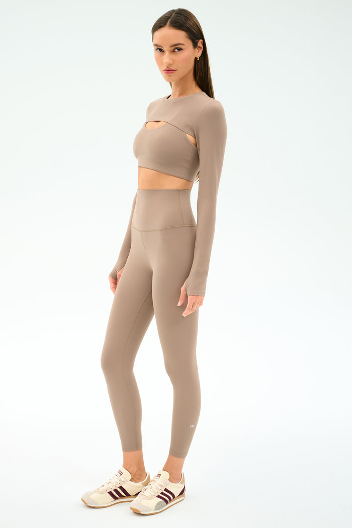 A woman stands in a taupe athletic set with long sleeves and high-waisted leggings, cream and maroon sneakers, and the SPLITS59 Amelia Airweight Lite Shrug - Latte—perfect for yoga or dance warmups—on a white background.