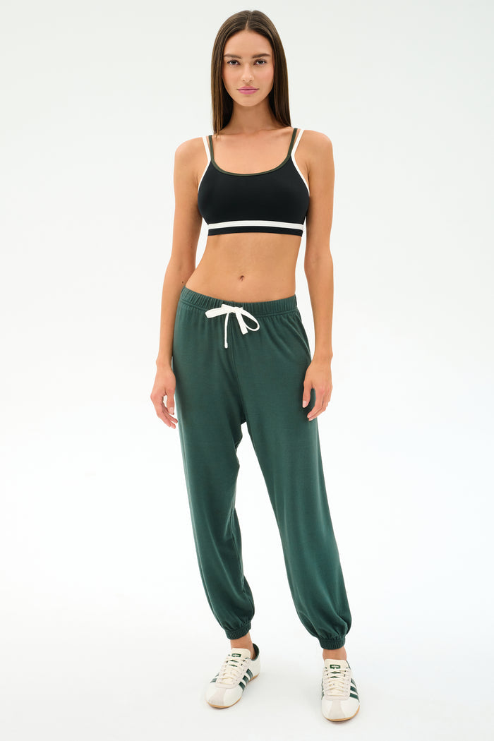 A woman faces forward in a black sports bra, SPLITS59's Andie Oversized Fleece Sweatpant in Military, and white sneakers—ideal for cool weather workouts—set against a plain white background.