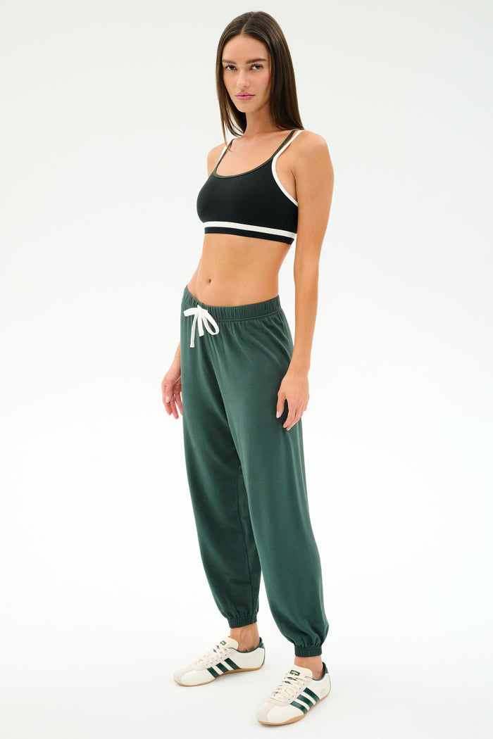 A woman stands against a plain background in a black sports bra, SPLITS59 Andie Oversized Fleece Sweatpant in Military, and white sneakers with green stripes—ideal for warm weather workouts.