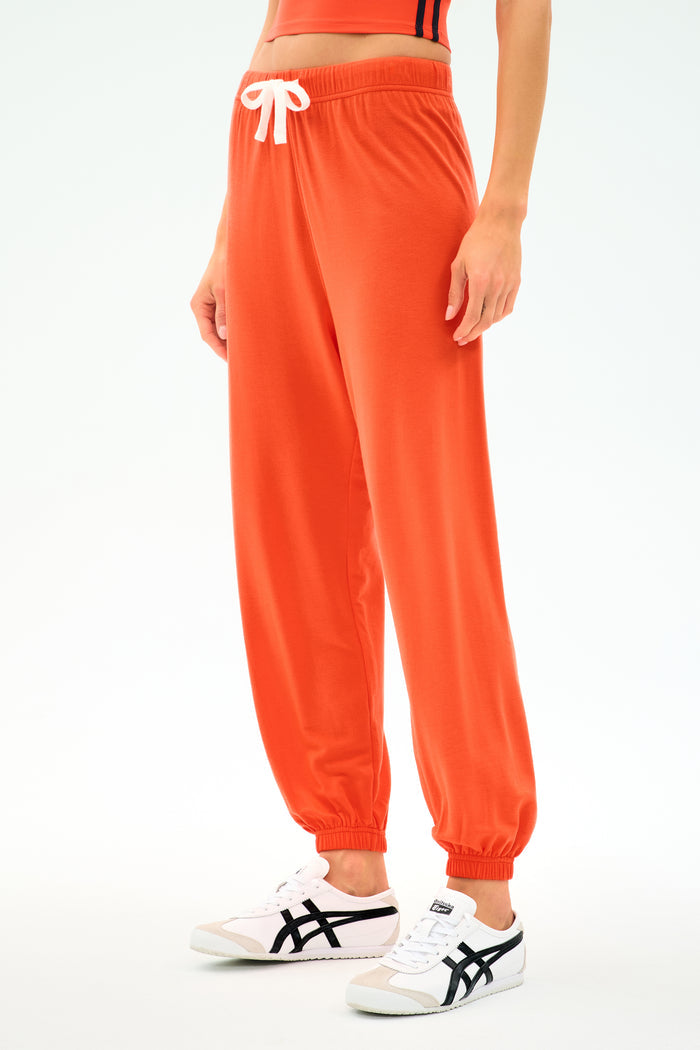 A person wearing SPLITS59's Andie Oversized Fleece Sweatpant in Valencia, an eye-catching orange, paired with white sneakers with black accents, stands against a plain light background.