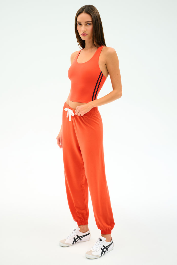A woman wears SPLITS59's Andie Oversized Fleece Sweatpant in Valencia—an orange, relaxed-fit pant with a white drawstring—paired with white sneakers featuring black stripes, standing against a plain light background.
