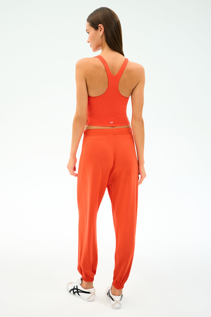 A woman faces away, wearing SPLITS59's Andie Oversized Fleece Sweatpant in Valencia with a matching orange sports bra and white sneakers against a plain white background.