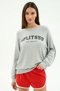 A woman stands against a plain white background wearing the SPLITS59 Andie Oversized Fleece Varsity Graphic Sweatshirt in Heather Grey/Splits59 Los Angeles and red athletic shorts.