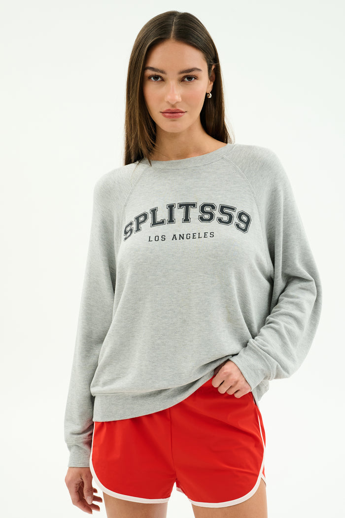 A woman stands against a plain white background wearing the SPLITS59 Andie Oversized Fleece Varsity Graphic Sweatshirt in Heather Grey/Splits59 Los Angeles and red athletic shorts.