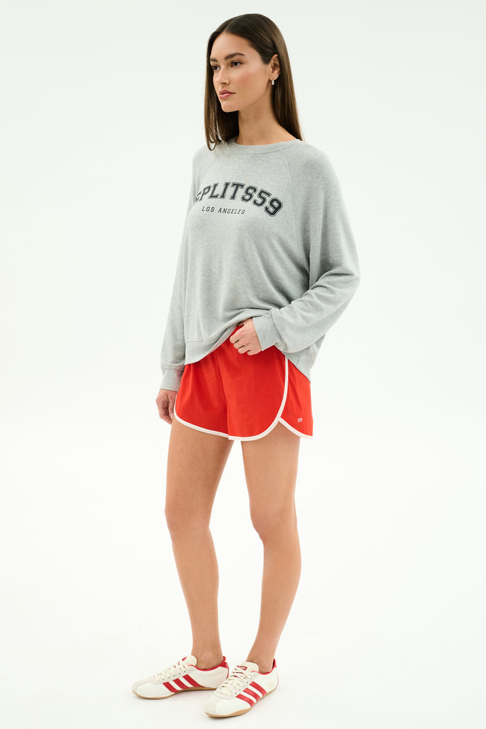 A woman wears the SPLITS59 Andie Oversized Fleece Varsity Graphic Sweatshirt in Heather Grey and red athletic shorts, styled with white sneakers. This cozy Los Angeles-made look features comfort and varsity flair.