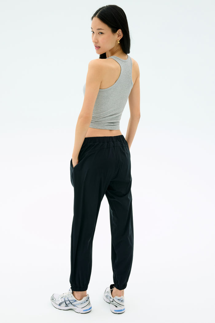 A woman stands with her back turned, wearing the Splits59 Ashby Rib Tank in Lt. Heather Grey, black jogger pants, and white sneakers, hands in pockets against a plain background.