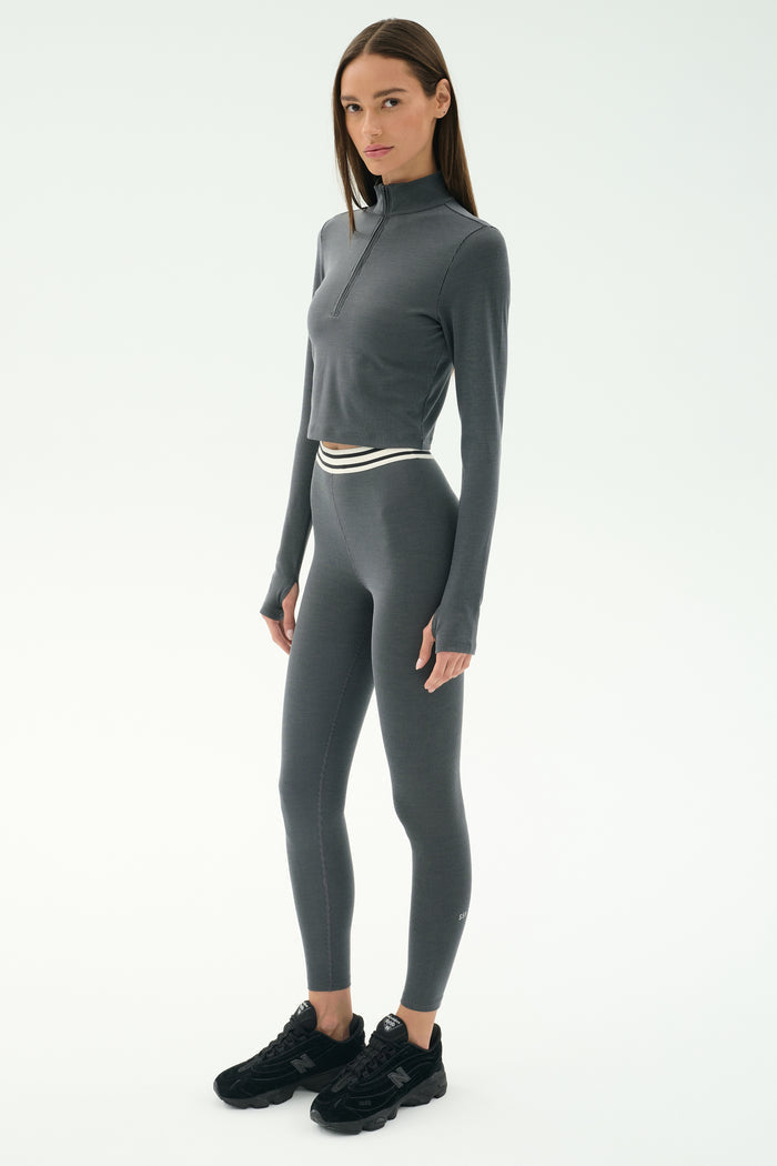 A woman wears the SPLITS59 Bailey Active Rib High Waist 7/8 leggings in Heather Grey with a fitted, long-sleeve top and black sneakers, standing against a plain white background.