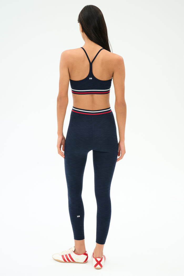 A woman faces away wearing the SPLITS59 Bailey High Waist Active Rib 7/8 leggings in Heather Indigo with matching sports bra, both featuring red, white, and navy stripes—ideal for the gym. She completes the look with white sneakers with red stripes.