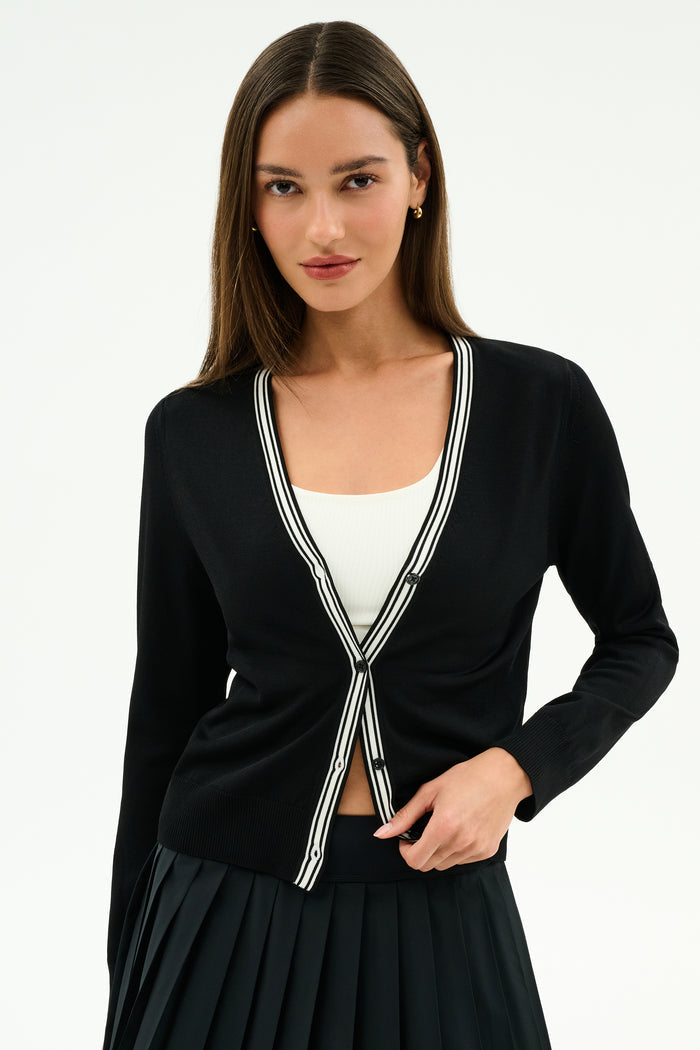 A woman models the SPLITS59 Bekah Knit Cardigan in Black/White—a soft rayon-nylon blend with white striped trim—over a white top and black pleated skirt, standing against a plain white background.