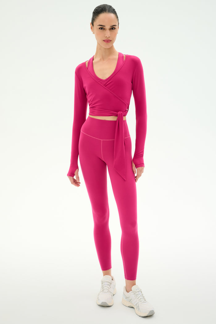 A woman stands against a plain background wearing the SPLITS59 Bella Airweight Lite Wrap in Sangria with matching high-waisted leggings and white sneakers.