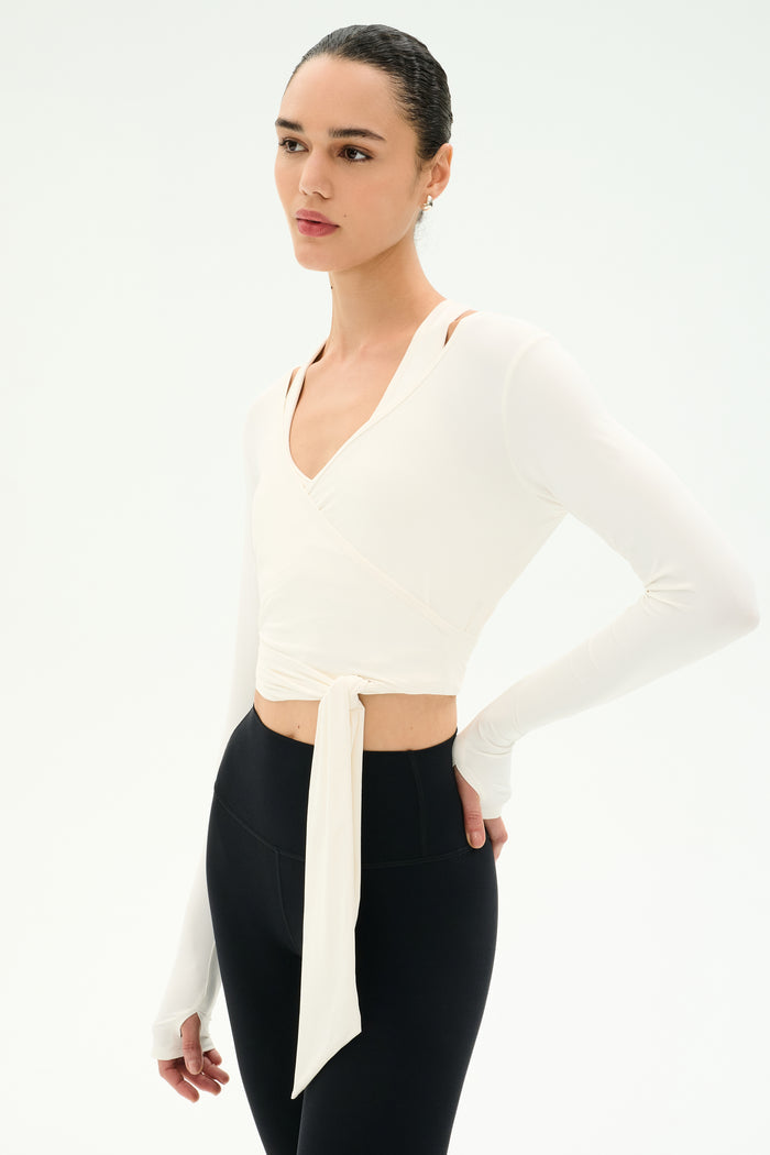 A person wearing the SPLITS59 Bella Airweight Lite Wrap in white stands against a plain white background, paired with black high-waisted nylon-spandex leggings.