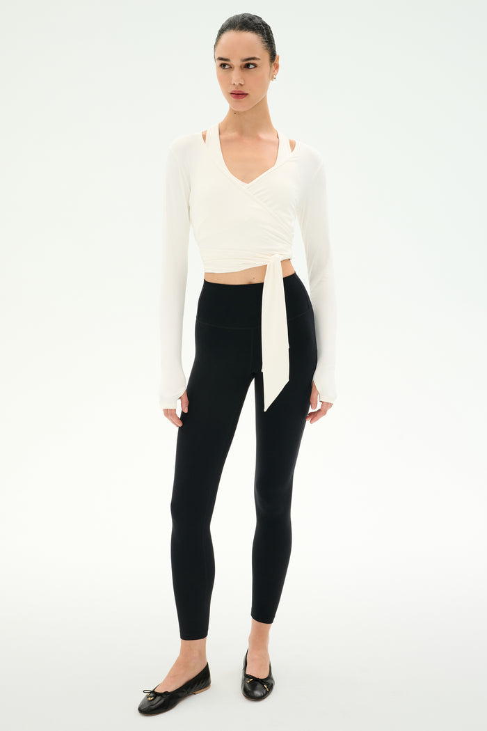 A woman wears the SPLITS59 Bella Airweight Lite Wrap in white, paired with black leggings and ballet flats, standing against a plain white background.